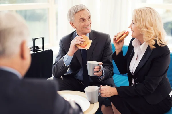 Business lunch in the airport lounge. An elderly man and an elderly woman in suits eat burgers. — Stock Photo, Image