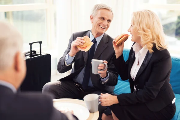 Almuerzo de negocios en la sala del aeropuerto. Un anciano y una anciana en traje comen hamburguesas . — Foto de Stock