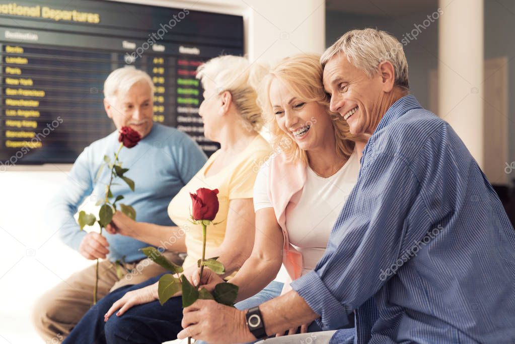 An elderly men gives roses to an elderly women. They are sitting on the couch. Elderly couple in airport.