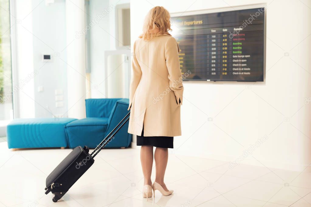 A woman is standing by the timetable at the airport. A woman is holding a suitcase.