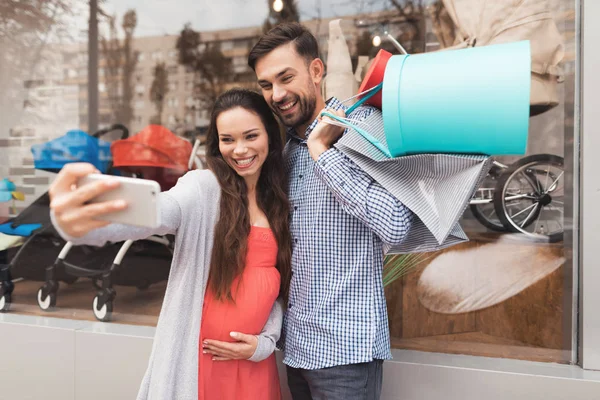 Una mujer embarazada con un hombre está haciendo una selfie cerca del escaparate con productos para niños. . — Foto de Stock