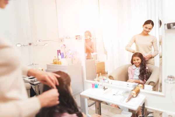 La peluquera peinando a una pequeña chica morena. La chica está sentada en la silla del salón de belleza . — Foto de Stock