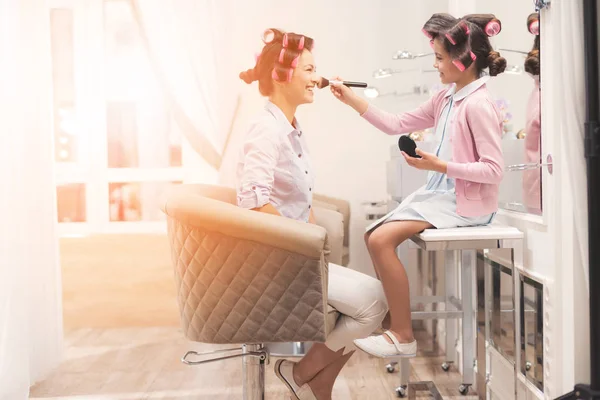 Moeder en dochter in de schoonheidssalon. Ze hebben krulspelden in hun haar. Meisje doet make-up aan moeder. — Stockfoto