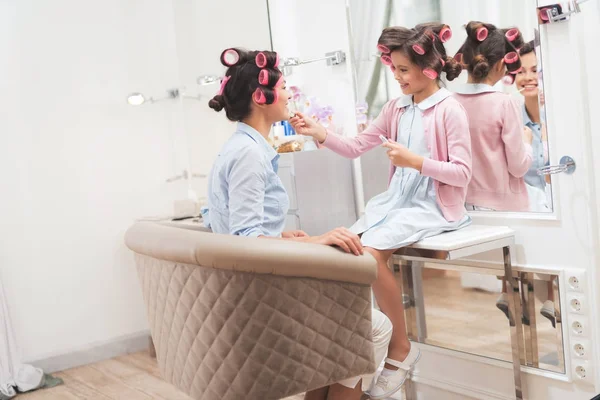 Mom and daughter in beauty salon. They have curlers in their hair. Little girl doing makeup to mom.