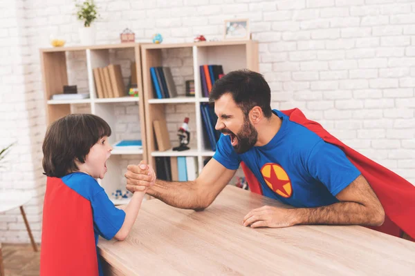 Father and son in suits of superheroes. They compete in armwrestling. They are measure their strength