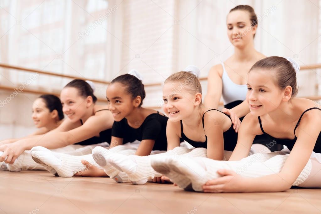 The trainer of the ballet school helps young ballerinas perform different choreographic exercises. They rehearse in the ballet class. The teacher communicates with the children.