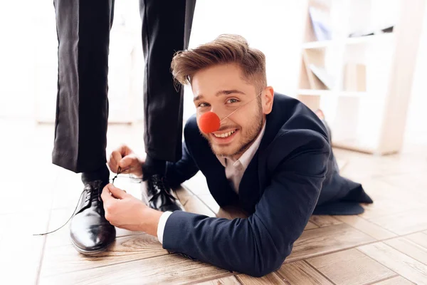Un uomo con il naso da clown sotto il tavolo per legare i lacci al suo collega d'affari. . — Foto Stock