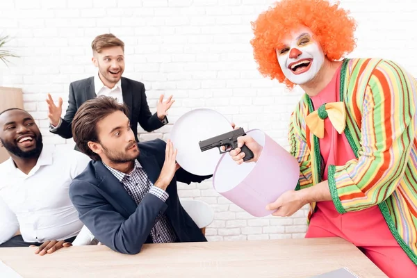 A man in a clown suit is aiming a gun at businessmen in a bright office. — Stock Photo, Image