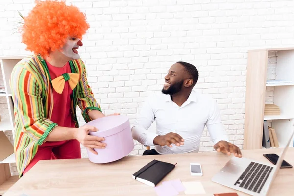 A man in a clown suit is holding a box with a surprise in his hand.