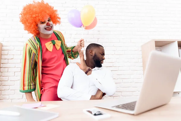 Człowiek w garniturze clown stoi obok czarny człowiek, który siedzi przy biurku. — Zdjęcie stockowe