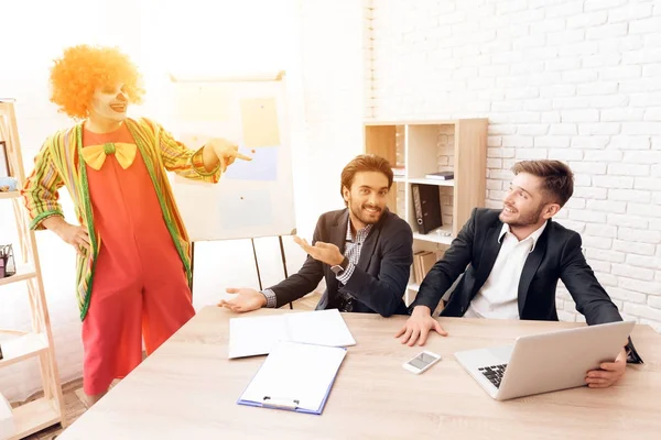 Человек в костюме клоуна стоит рядом с мужчинами в деловых костюмах, которые сидят за столом . — стоковое фото