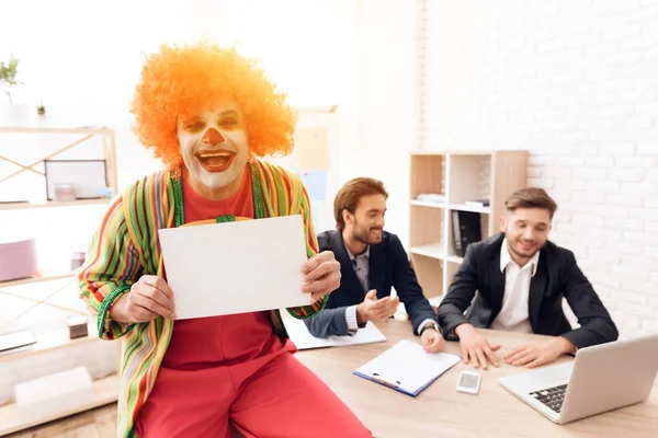 Człowiek w garniturze clown stoi obok mężczyzn w garniturach, którzy siedzą przy biurku. — Zdjęcie stockowe