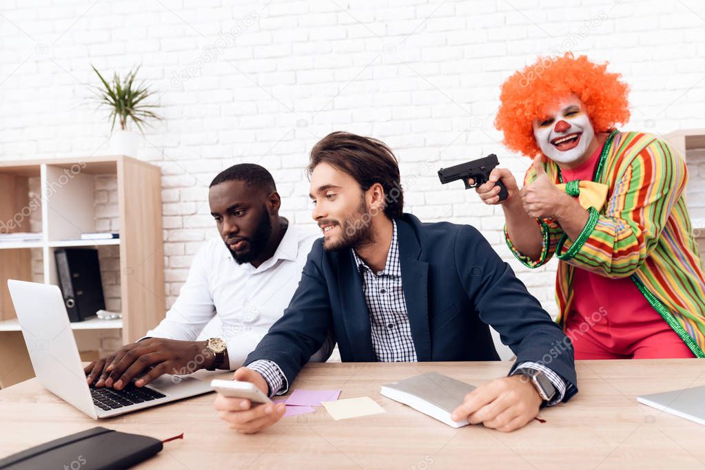 A man in a clown suit is aiming a gun at businessmen in a bright office.