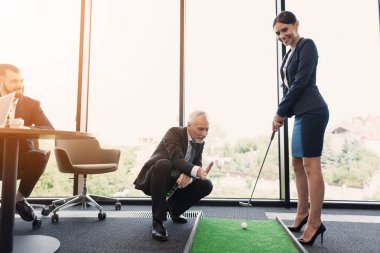 Siyah takım elbiseli bir kadın ofiste golf oynuyor. İş takım elbiseli yaşlı bir adam ona yardımcı olur