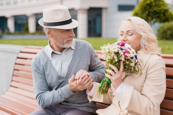 Dos pensionistas están sentados en un banco en el callejón. El anciano le dio flores a la mujer. Él sostiene su mano — Foto de Stock