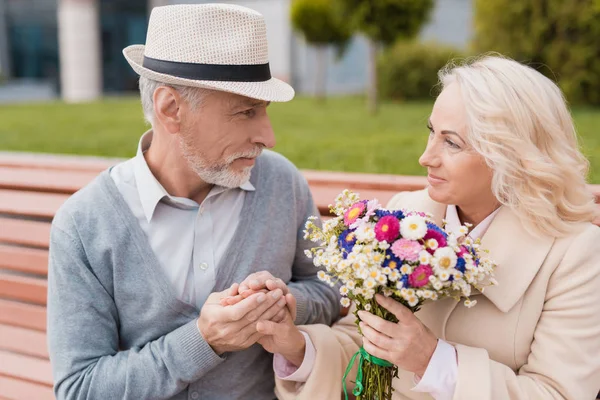 Dos pensionistas están sentados en un banco en el callejón. El anciano le dio flores a la mujer. Él sostiene su mano — Foto de Stock