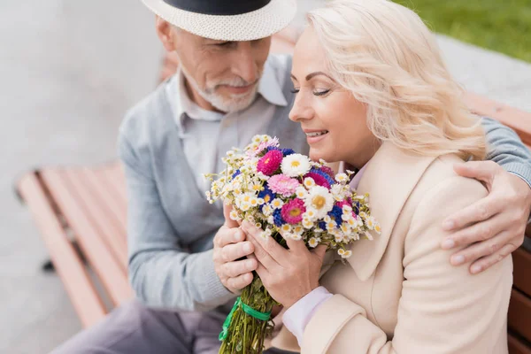 Δύο συνταξιούχοι κάθονται σε ένα παγκάκι στο δρομάκι. Ο μεσήλικας άνδρας έδωσε τα λουλούδια της γυναίκας. Κρατάει το χέρι — Φωτογραφία Αρχείου