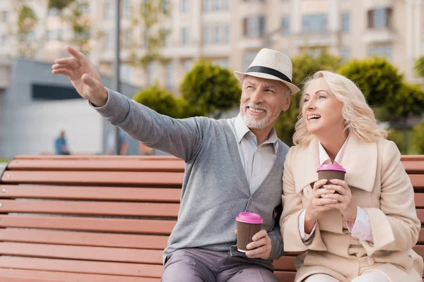 Dos pensionistas están sentados en un banco con un vaso de café en sus manos. El hombre le muestra a la mujer algo por delante — Foto de Stock