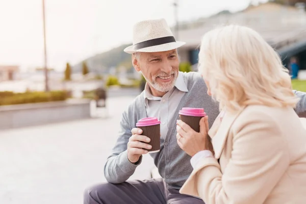 Dos pensionistas están sentados en un banco con un vaso de café en sus manos. Se miran con ternura. — Foto de Stock