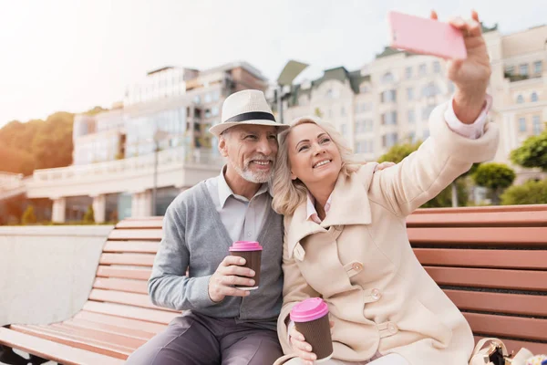 Dos pensionistas están sentados en un banco con un vaso de café en sus manos. Hacen selfies en el teléfono inteligente de una mujer — Foto de Stock