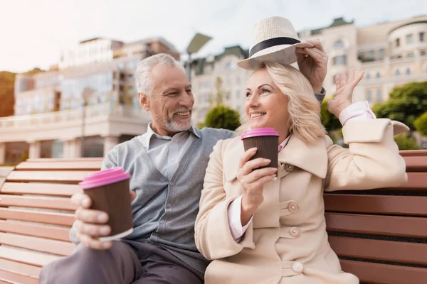 Dos pensionistas están sentados en un banco con un vaso de café en sus manos. Se sientan abrazando — Foto de Stock