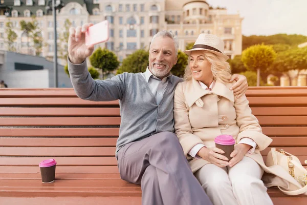 Dos pensionistas están sentados en un banco con un vaso de café en sus manos. Se sientan abrazando y haciendo selfies — Foto de Stock