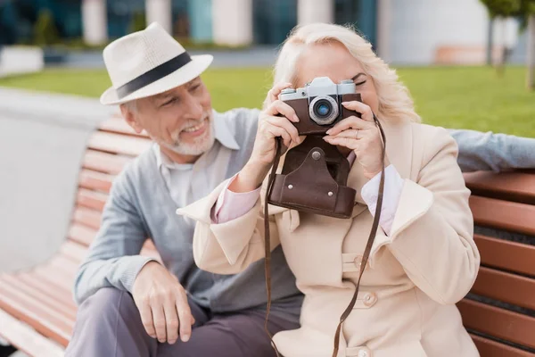 Двое пенсионеров сидят на скамейке и фотографируют на старую кинокамеру. Они улыбаются. — стоковое фото