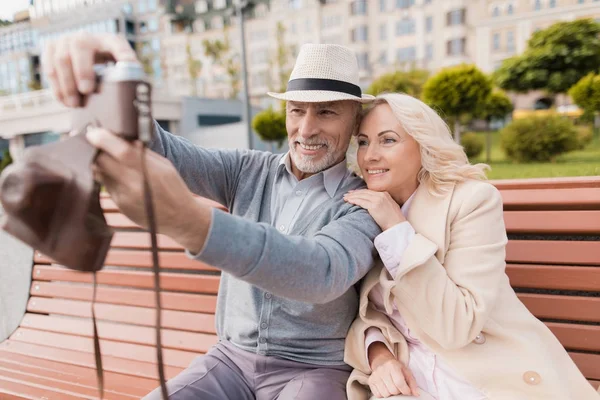 Un par de jubilados están sentados en un banco en el centro comercial. Hacen selfie en una vieja cámara de cine — Foto de Stock