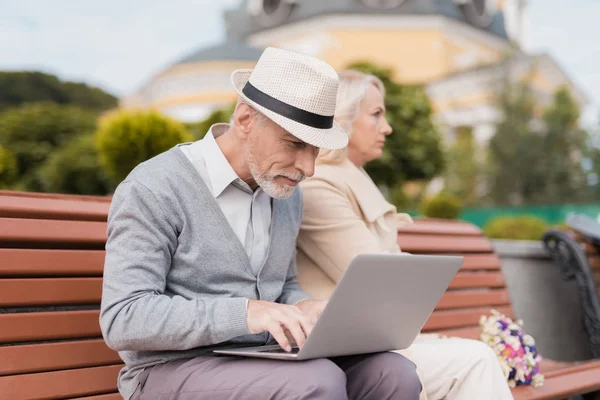 A couple of retired people are sitting on a bench in the mall. A man is working on a laptop, a woman is offended at him