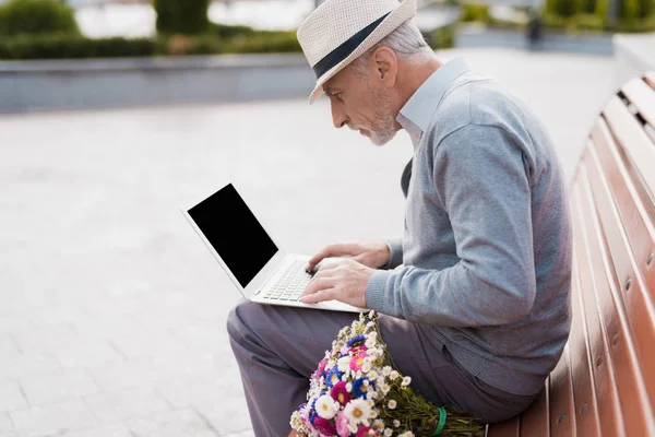 The pensioner sits on the bench and works behind the laptop. He sits on a bench in the alley.