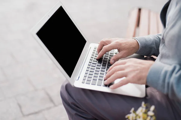 Pensionären sitter på bänken och fungerar bakom den bärbara datorn. Han sitter på en bänk i gränden. — Stockfoto