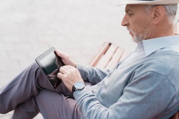 Un pensionista se sienta en un banco en una hermosa plaza. Tiene una tableta gris en sus manos. Está leyendo algo. — Foto de Stock