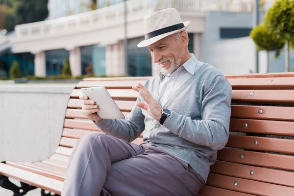 En pensionär som sitter på en bänk på ett vackert torg. Han har en grå tablett i händerna. Han kommunicerar genom video med någon — Stockfoto