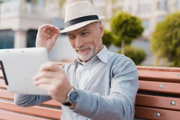 En pensionär som sitter på en bänk på ett vackert torg. Han har en grå tablett i händerna. Han kommunicerar genom video med någon — Stockfoto