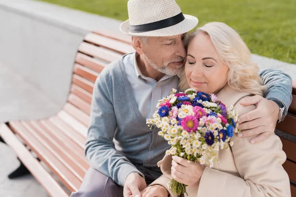 Dos pensionistas están sentados en un banco en la plaza. Se abrazan. Una mujer tiene un ramo de flores en sus manos. — Foto de Stock