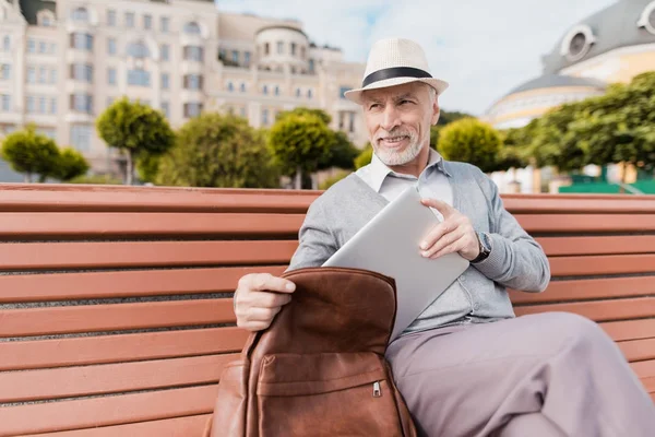En pensionär som sitter på en bänk på ett vackert torg. Han tar ut en grå tablett från sin portfölj — Stockfoto