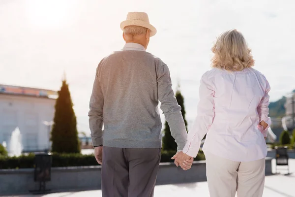 Una pareja de ancianos está caminando tomados de la mano. Caminan a lo largo de una hermosa plaza — Foto de Stock