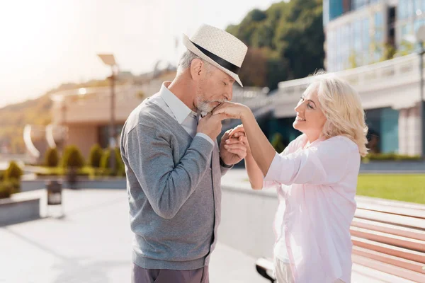 An elderly couple is walking in the square. The man kisses the woman\'s hand. She smiles