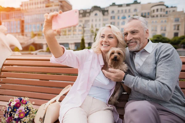 Um casal de idosos está andando na praça com seu cachorrinho. Fazem uma selfie com um cão. — Fotografia de Stock
