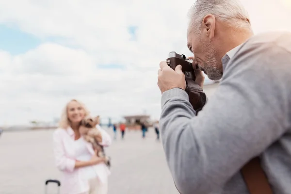 Una pareja de ancianos está caminando. Una mujer posa con un perro en sus brazos, un hombre toma fotos de ella en una cámara de cine — Foto de Stock