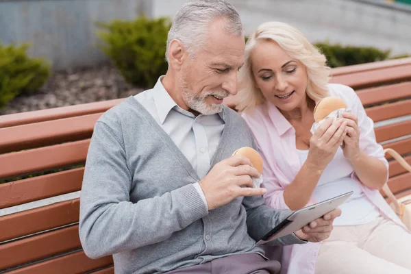 An elderly couple is resting sitting on a bench in the square. They eat burgers and look at something on the tablet