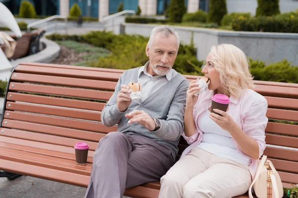 An elderly couple is resting sitting on a bench in the square. They eat burgers and drink coffee