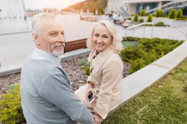 Una pareja de ancianos está sentada en el borde de un macizo de flores. Se sonríen. — Foto de Stock