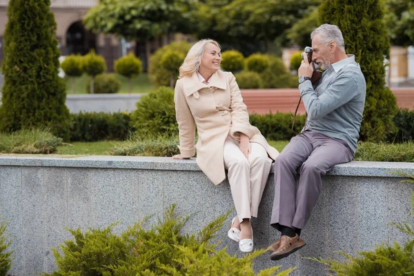 Una pareja de ancianos está sentada en el borde de un macizo de flores. Se toman fotos el uno al otro en una cámara de cine vintage —  Fotos de Stock
