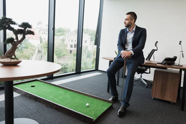 A man in a strict business suit is sitting on the edge of a table with a golf cake. Near a golf mat in the office