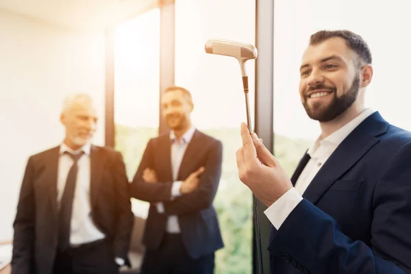 Un homme en costume d'affaires noir respectable examine un club de golf. À propos des collègues jouant au golf au bureau — Photo