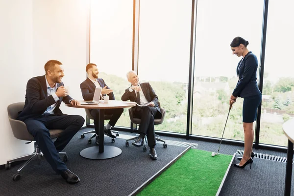Tres hombres se sientan en una mesa y ven a una mujer en un traje de negocios jugando mini golf en la oficina — Foto de Stock