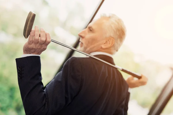 Een bejaarde respectabel man die zich voordeed in zijn kantoor met een golfclub. Hij zit op het bureaublad achter de laptop — Stockfoto
