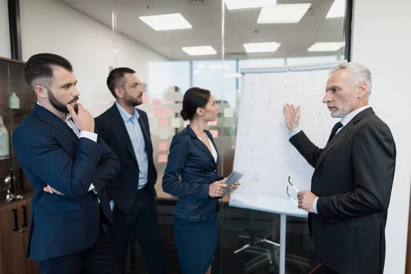 Three employees and their director are next to the rack with the schedule. They carefully consider it. — Stock Photo, Image