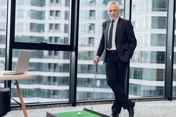 Un hombre respetable de edad avanzada en un estricto traje de negocios posando en la oficina jugando un mini juego de golf — Foto de Stock